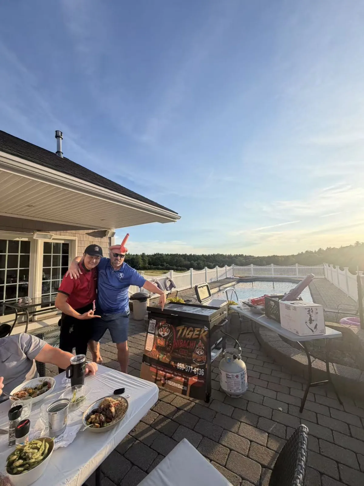Two hosts standing behind the teppanyaki grill at a lakeside hibachi party at dusk in Rhode Island