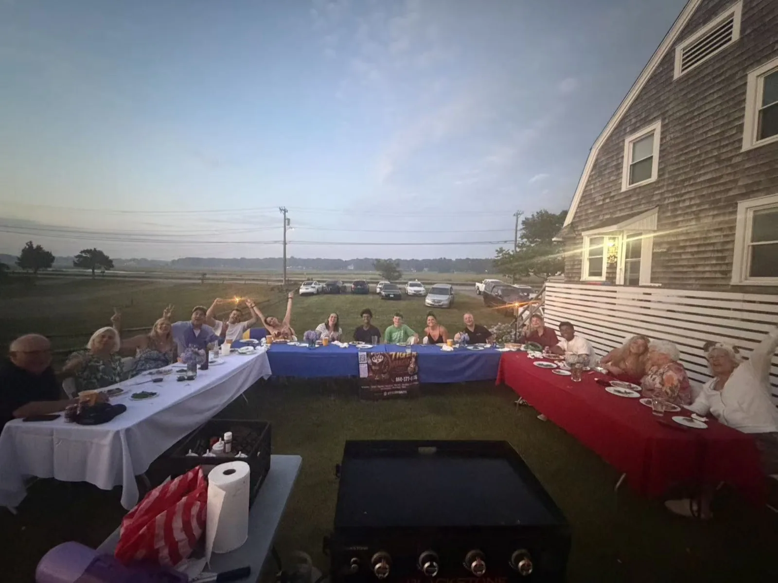 Long dinner table set for a waterfront hibachi party at dusk with a shingle-style house in Rhode Island